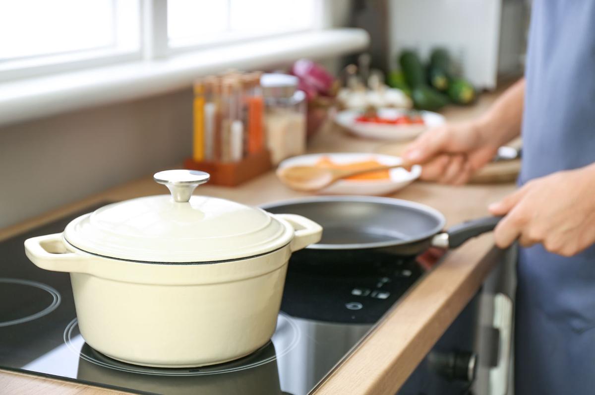 A person in a blue shirt cooking in a kitchen with a cream-colored pot on the stove and a frying pan being handled. Ingredients and spices are visible in the background.